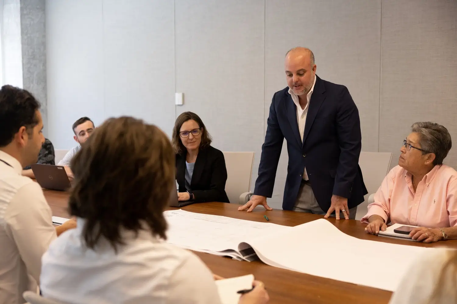 A group of professionals gathers around a conference table to review large project drawings, with one man standing and speaking to the team.
